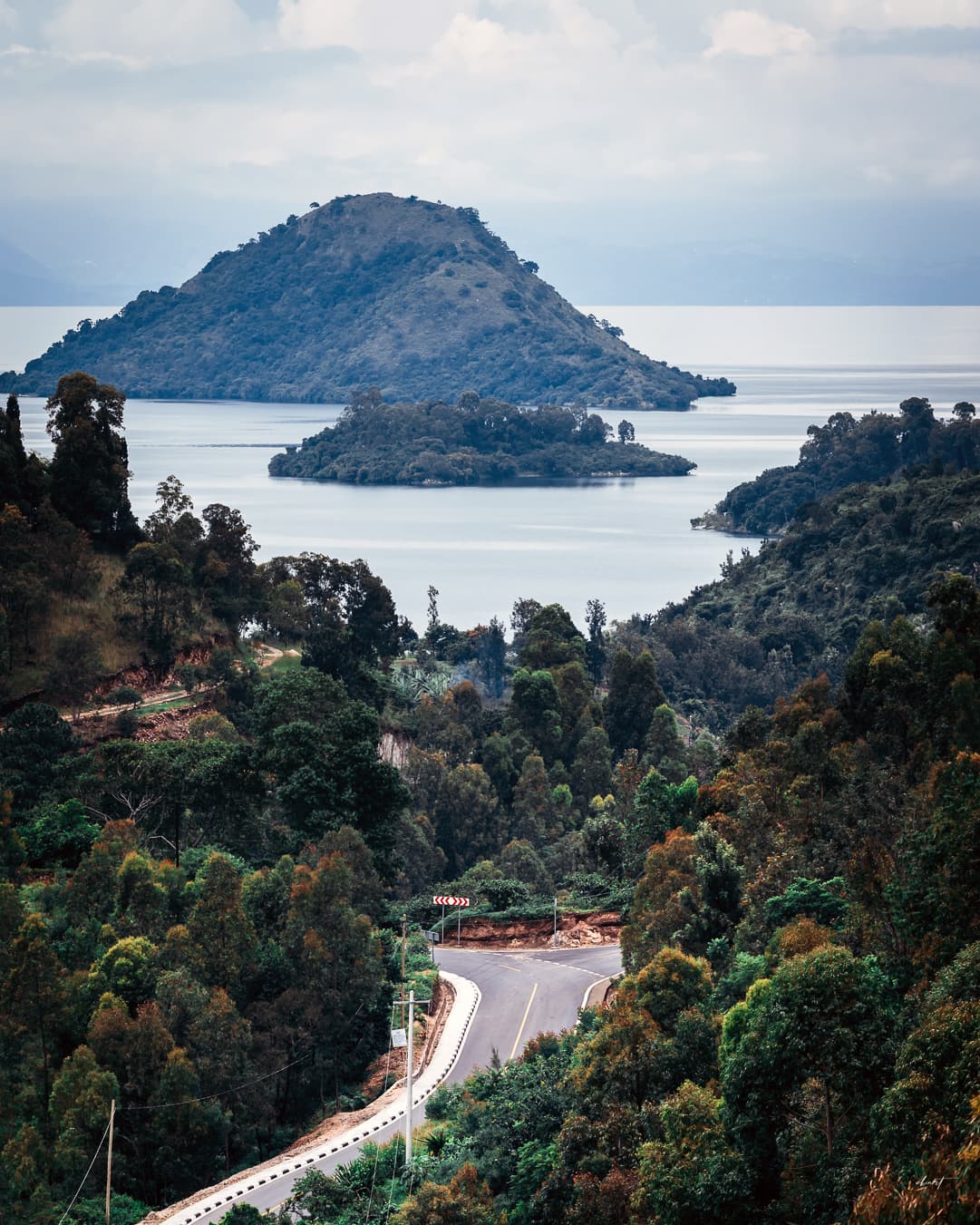 A new tarmac road winding down through forested hills toward a lake in Rwanda